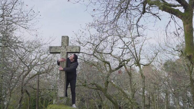 Uomo adulto che tocca una grande croce di pietra in un tranquillo parco all'aperto in una giornata nuvolosa.