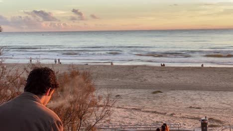 Die-Silhouette-Eines-Mannes-Sitzt-Auf-Einer-Mauer-Mit-Blick-Auf-Den-Strand,-Raucht-Eine-Elektronische-Zigarette-Und-Blickt-Nachdenklich-Auf-Den-Sonnenaufgang