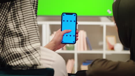 woman holding a phone with a blue screen, in front of a green screen tv, with another woman in background