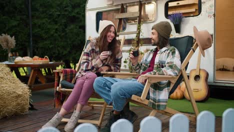 een gelukkig brunette meisje in een hoed en een geruite hemd samen met haar brunette vriend in een groen geruite shirt drinken thee, zitten op stoelen en praten bij een trailer tijdens hun picknick in een kamp buiten de stad in de zomer