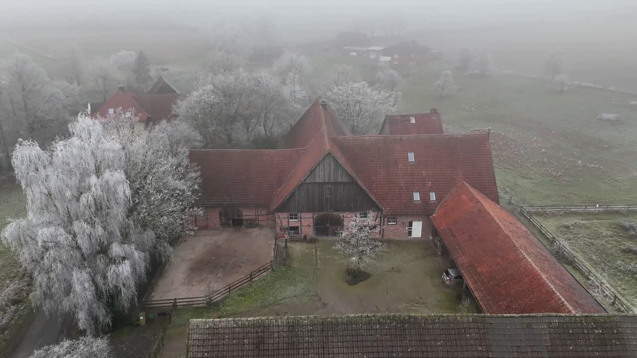 Farm house with stable and frozen fields during cold and foggy winter day. Aerial top down flyover shot. Historic and ancient farm buildings in German countryside.