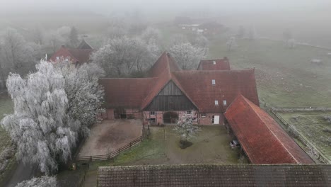 farm house with stable and frozen fields during cold and foggy winter day. aerial top down flyover shot. historic and ancient farm buildings in german countryside.