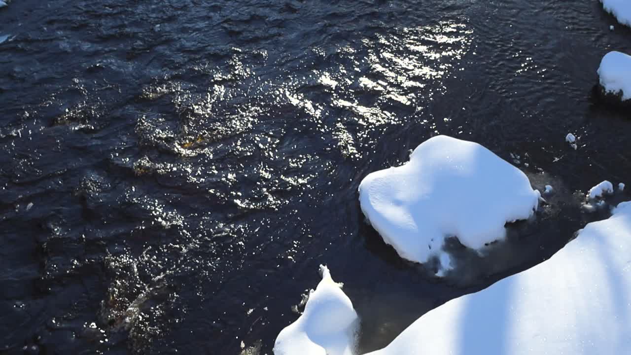 Top down angle footage of dark cold river water flowing in slow motion during a winter sunny day, with sunshine reflecting on the water ripples and waves. Snow covered rocks in the water visible also.