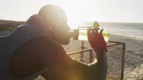 Hombre-Afroamericano-Bebiendo-Agua,-Tomando-Un-Descanso-En-El-Ejercicio-Al-Aire-Libre-Junto-Al-Mar