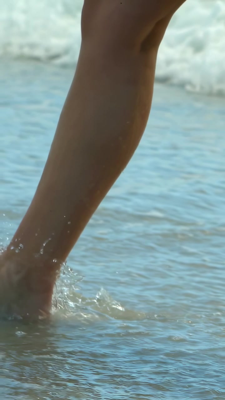 Video verticale: Donna che solleva il piede e cammina sulla sabbia bagnata e sulla risacca bassa, indossando pantaloncini.