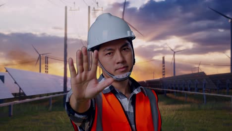 close up of asian male engineer with safety helmet disapproving with no hand sign while standing with solar panel and wind turbines