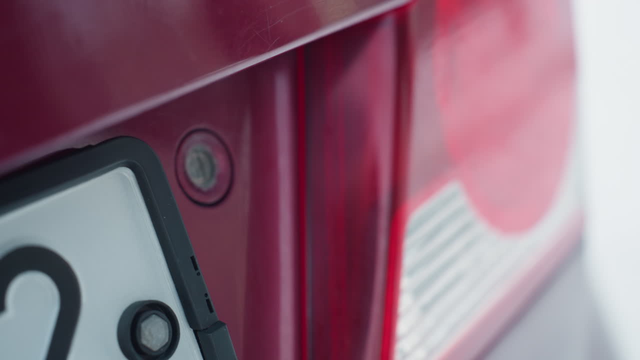 close up of car bonnet as person inserts key into lock and turns to secure hood latch under cold outdoor lighting, glossy paint reflects sky, blurred trees and building in background