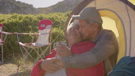 Happy-senior-biracial-couple-sitting-at-tent-in-mountains-and-embracing-on-sunny-day,-in-slow-motion