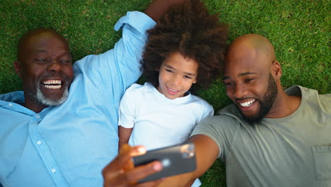 Draufsicht-Einer-Männlichen-Mehrgenerationenfamilie,-Die-Im-Gras-Liegt-Und-Ein-Selfie-Mit-Dem-Mobiltelefon-Macht