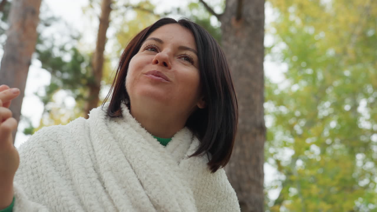 Mulher enrolada em um cobertor ao ar livre, comendo um lanche em uma aconchegante manhã de outono em uma floresta de pinheiros com manta de tricô grossa e suéter verde, mulher assando marshmallow em uma panela de acampamento, vapor subindo, sorriso gentil