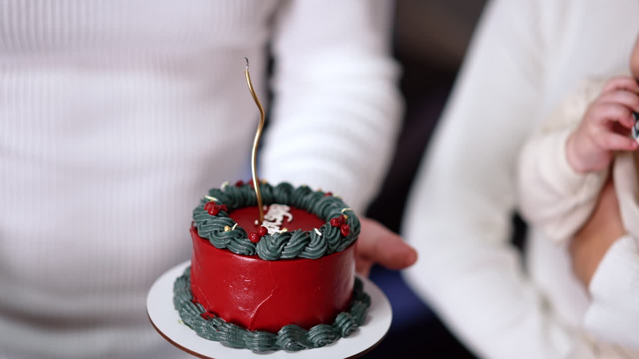 Unrecognized man holds a little red cake near the baby. Man lights the thin curved candle on the cake. Close up.
