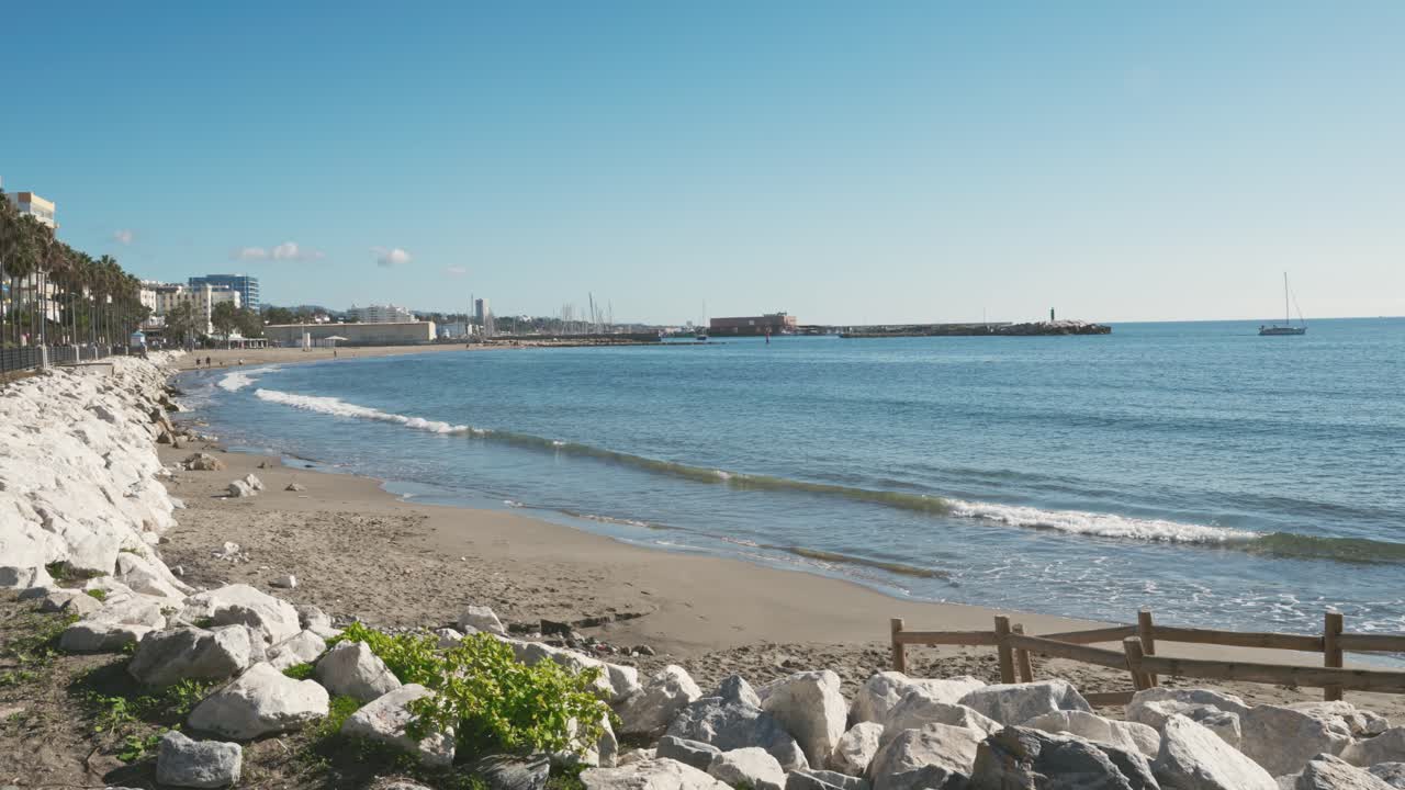 Ombak tenang di sepanjang tepi laut Marbella dengan Pantai La Bajadilla dan pelabuhan perikanan, Costa del Sol, Spanyol Selatan, di bawah langit biru cerah, menangkap pesona Mediterania