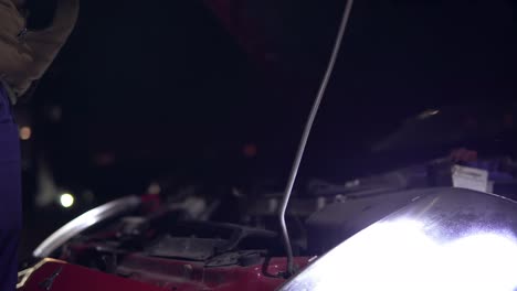 hombre mirando el motor de un coche averiado con una linterna por la noche. luces encendidas