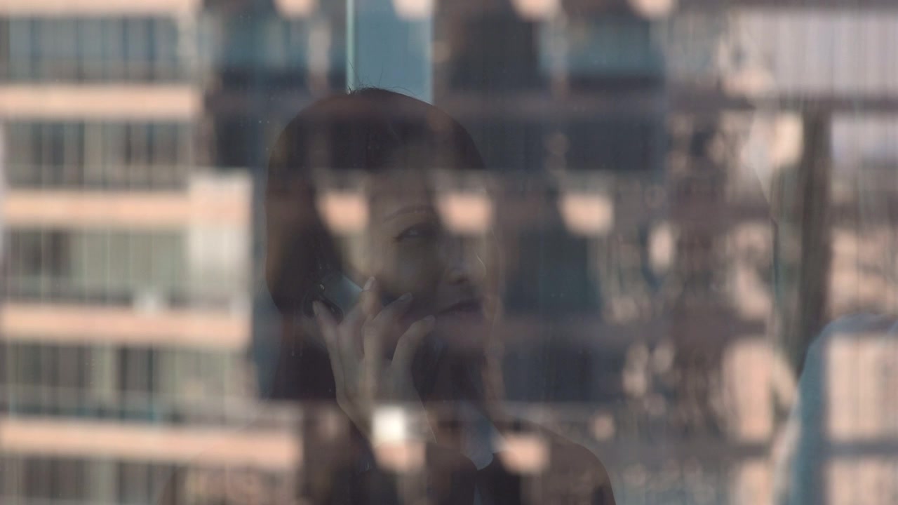 Duas empresárias no escritório falando ao celular e fechando negócios. A imagem é capturada através da janela, mostrando o reflexo dos prédios de escritórios ao redor na cidade.