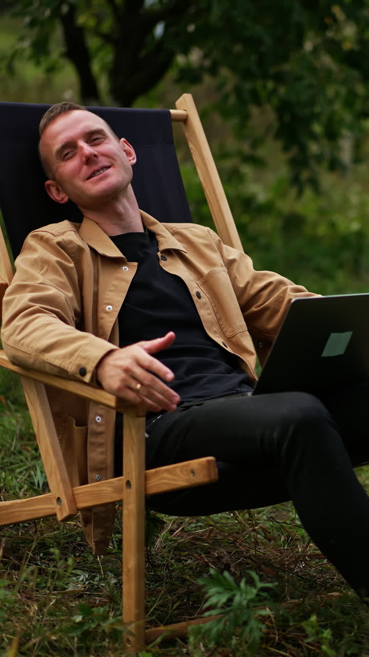 Happy relaxed man laid back in the garden chair. Male freelancer working remotely in the autumn garden. Man speaking on video chat on his laptop. Vertical video