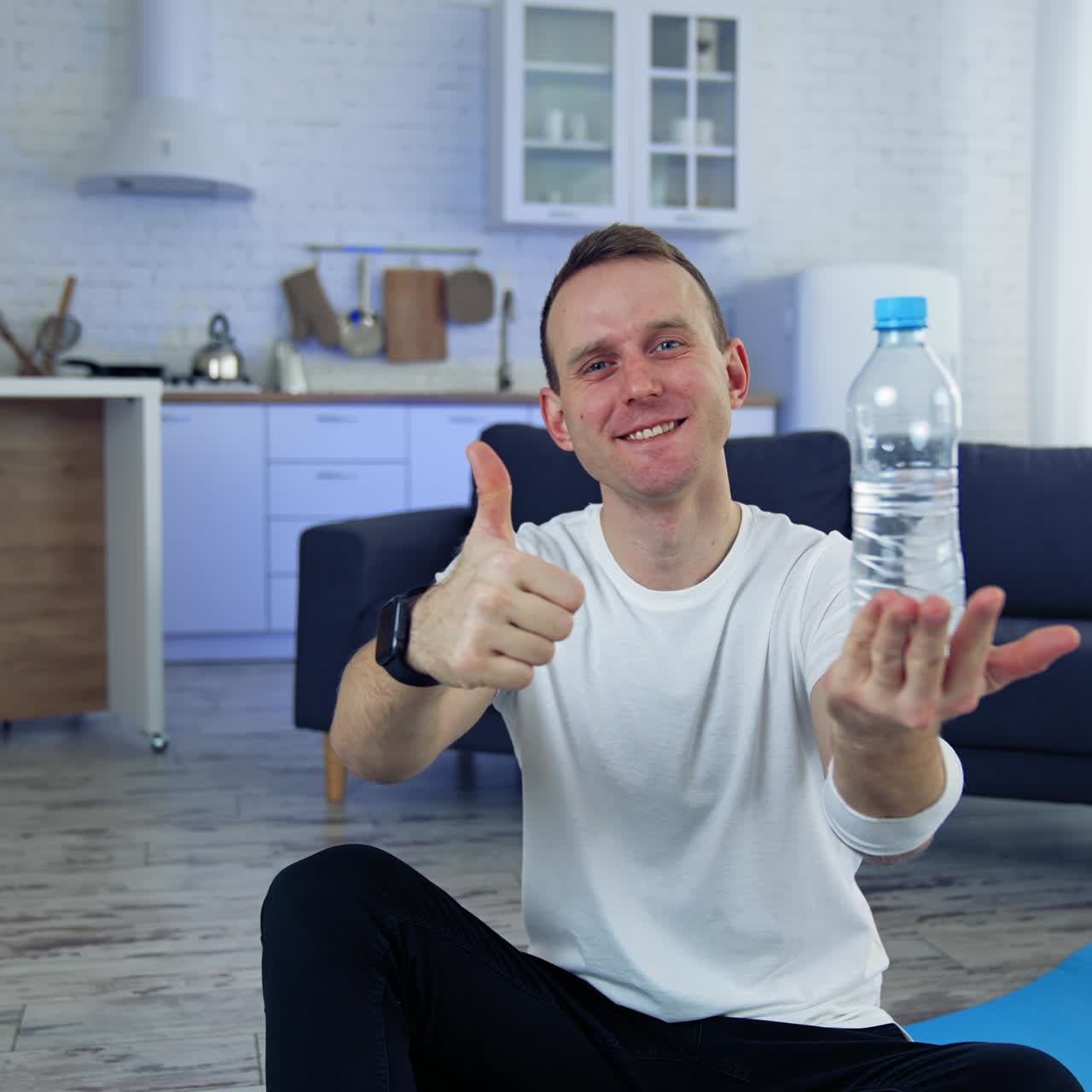 Man with a bottle of water at home. Young man in white t-shirt sitting on a floor and showing a bottle after doing fitness exercises
