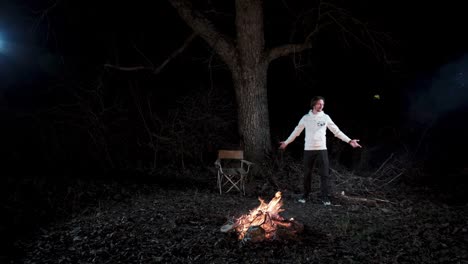 Static shot of a man standing along by a campfire in the forest