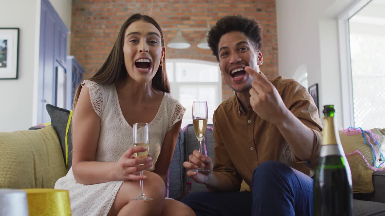 feliz pareja biracial bebiendo champán en casa haciendo una celebración llamada de video portátil y riendo