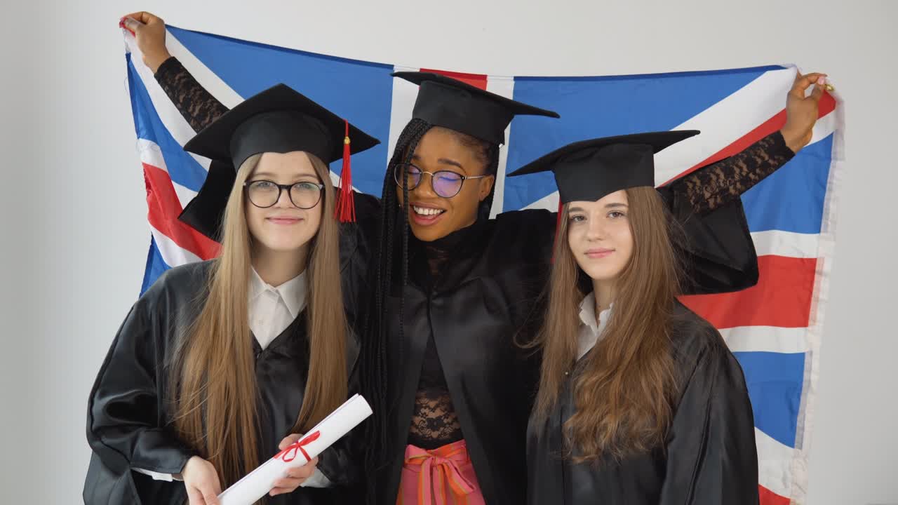 tres jóvenes graduadas felices de diferentes nacionalidades están juntas uno al lado del otro con diplomas en las manos y con la bandera británica detrás de ellas. vista de cerca sobre fondo blanco