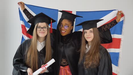 three young happy graduate women of different nationalities stand together side by side with diplomas in their hands and with british flag behind them. close up view on white background