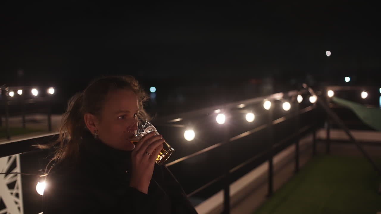 Donna che beve birra su una terrazza panoramica di notte con luci a stringa