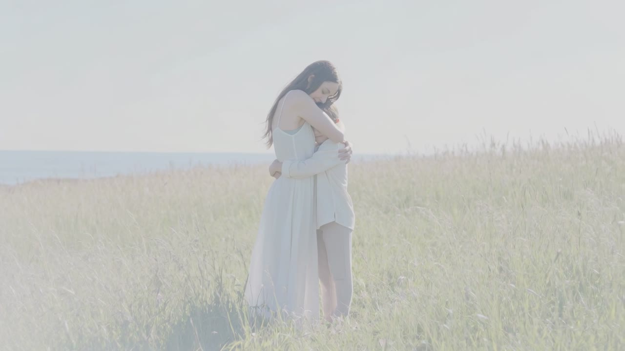 Duas mulheres em pé se abraçando em um prado ensolarado à beira-mar, com a brisa movendo a grama e os vestidos, para anúncios de romance.