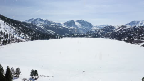 Frozen-lake-with-snowy-mountain-range-in-distance-during-heavy-snowfall,-aerial-drone-fly-forward-view