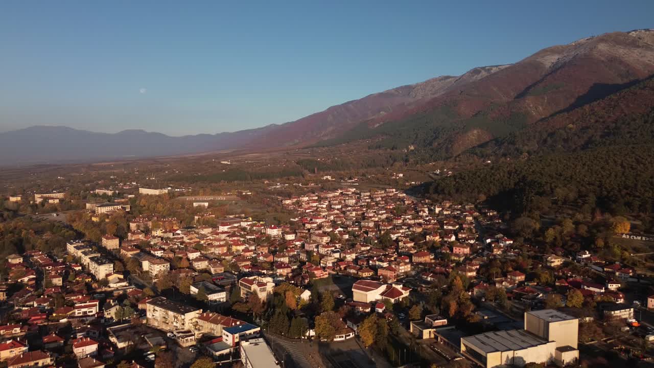 Enchanting morning drone footage of Sopot, Bulgaria, bathed in golden hour light. The majestic Balkan Mountains rise on the right, while the moon lingers in the blue sky, adding a touch of serenity