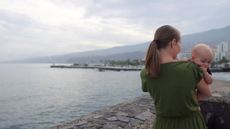 with her baby son, a young mother takes a leisurely walk by the ocean on a historic european square, observing the waves and smiling in contentment