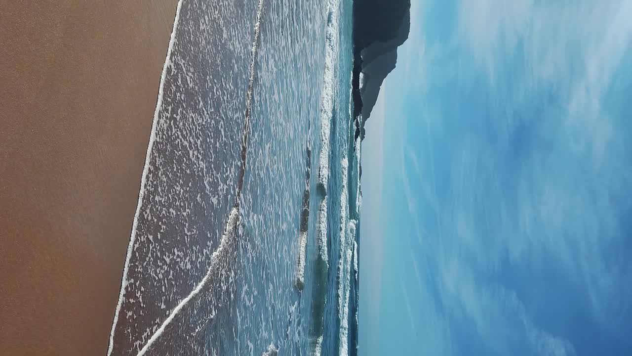 Zambujeira do Mar sulla costa con onde oceaniche, scogliere e dune di sabbia ricoperte da vegetazione verde e foglie rosse di fico degli Ottentotti, in una giornata di sole con cielo azzurro limpido