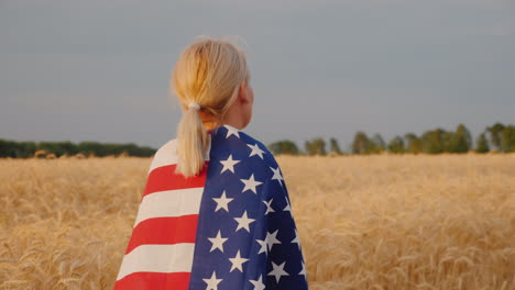 Eine-Junge-Frau-Mit-Der-Us-flagge-Auf-Ihren-Schultern-Steht-In-Einem-Feld-Aus-Reifem-Weizen-4k-Video