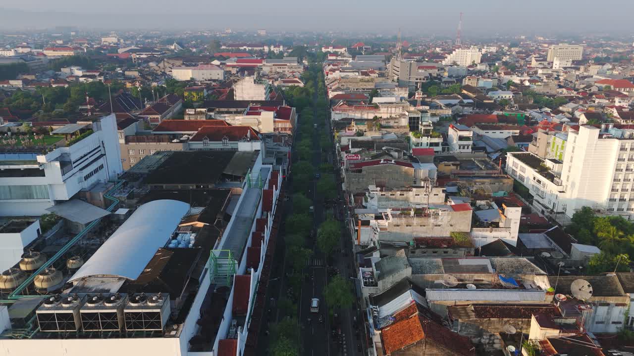 Um drone avança ao longo do famoso bulevar comercial com a luz da manhã, cercado por prédios urbanos, uma avenida central arborizada e tráfego tranquilo ao nascer do sol em Yogyakarta