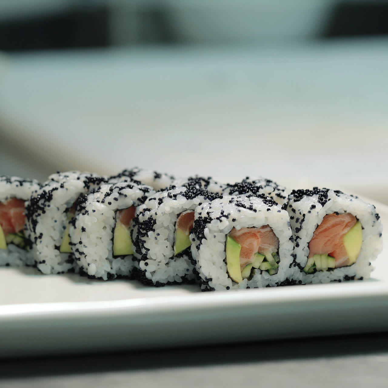 Käsineisiin pukeutunut sushikokki asettelee rullia koristellulle lautaselle sushibaarissa. Ammattikokki valmistaa rullia asiakkaalle ammattikeittiössä. Neliövideo.