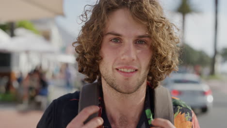 close up portrait of happy young man smiling enjoying warm summer vacation in busy urban beachfront wearing backpack