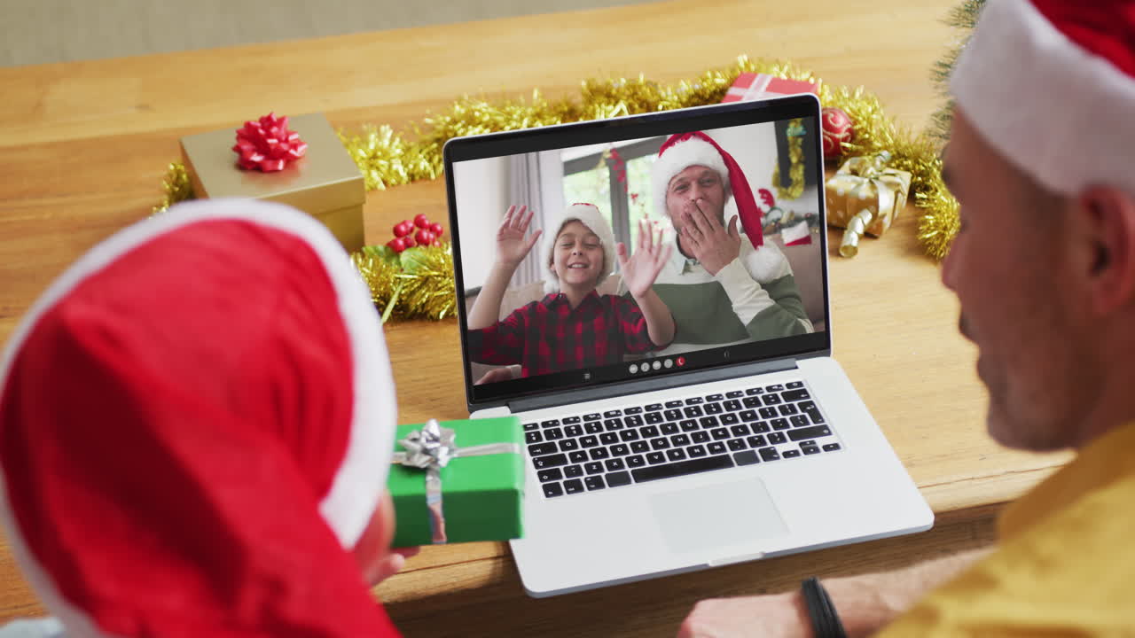 padre y hijo caucásicos con sombreros de santa usando una computadora portátil para una videollamada de navidad con la familia en la pantalla