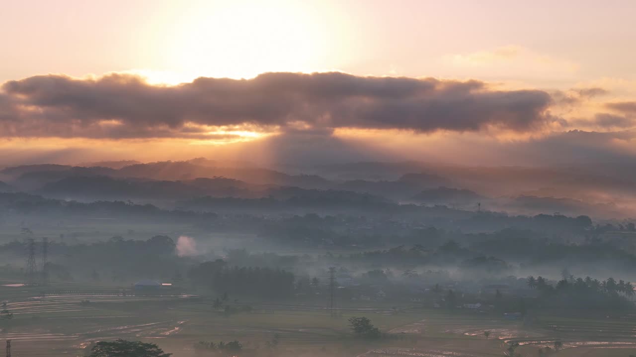 Aerial view of a peaceful countryside at dawn, with golden sunlight streaming through thick clouds and soft morning mist blanketing the landscape. Tropical rural landscape