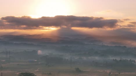 aerial view of a peaceful countryside at dawn, with golden sunlight streaming through thick clouds and soft morning mist blanketing the landscape. tropical rural landscape