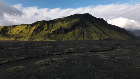 Aerial-tracking-shot-of-a-car-on-a-black-volcanic-road-with-a-mountain-in-the-background,-on-a-sunny-day-with-some-clouds