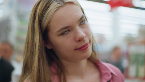 lady in pink dress inside a well-lit mall makes a funny facial expression while blinking her eyes, behind her, a man can be seen working, slightly blurred, the background shows a bright, lively mall