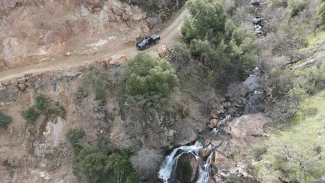 Aerial-drone-shot-of-a-black-pickup-truck-driving-past-a-waterfall