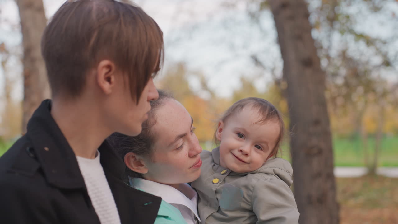 Giovane famiglia bianca che condivide un momento giocoso al parco, la madre culla un bambino sorridente mentre il padre si china vicino, luminoso sfondo autunnale, primo piano spontaneo, legame gioioso, baci delicati e risate, atmosfera accogliente.