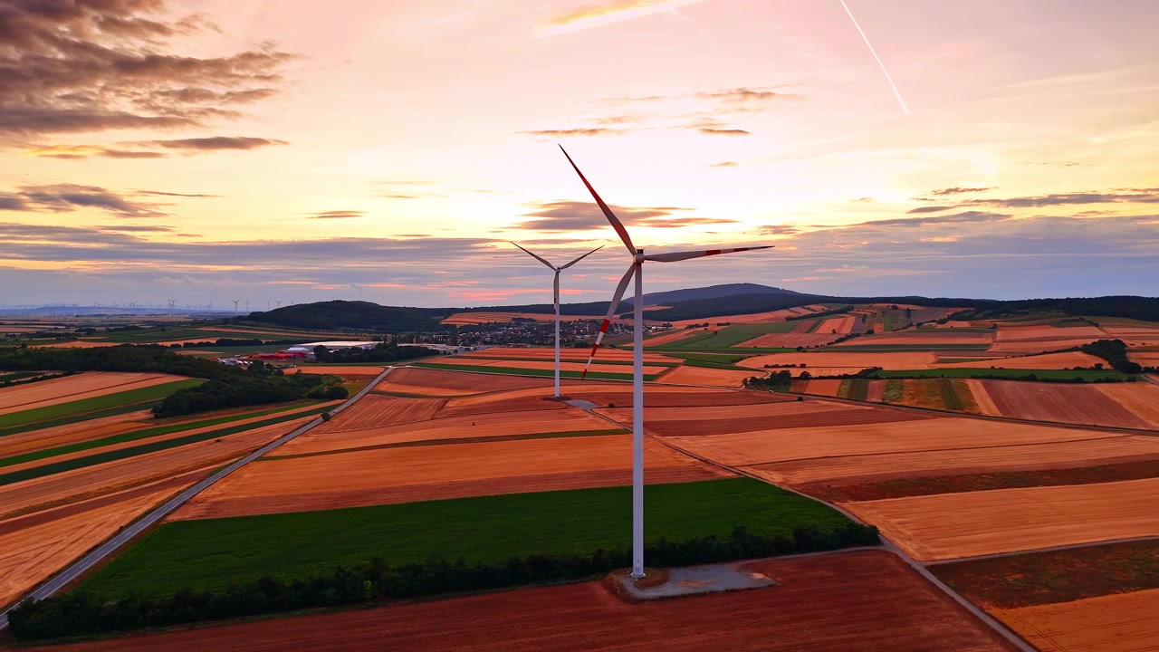 To vindmøller over geometrisk landbrugsland under orange solnedgang. Forhøjet udsigt viser hvide tårne med røde vingemarkeringer, landeveje, bakker og en baggrundsbelyst himmel