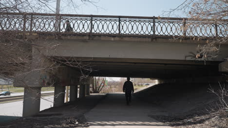 close view of a pedestrian walking under a bridge with decorative iron railings, it captures urban infrastructure, bare trees, and graffiti