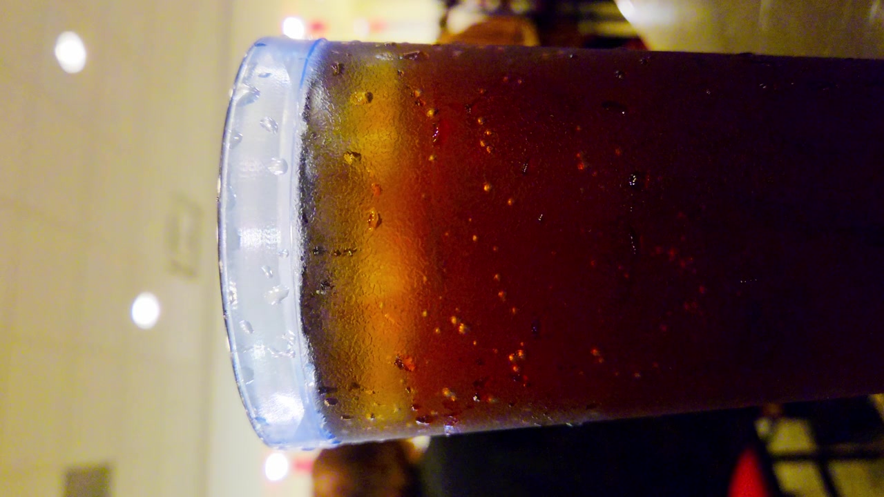 Primo piano di un bicchiere di bevanda ghiacciata con goccioline di condensa, in primo piano, con persone sfocate (effetto bokeh) sullo sfondo, di spalle. La scena è ambientata in un ristorante.