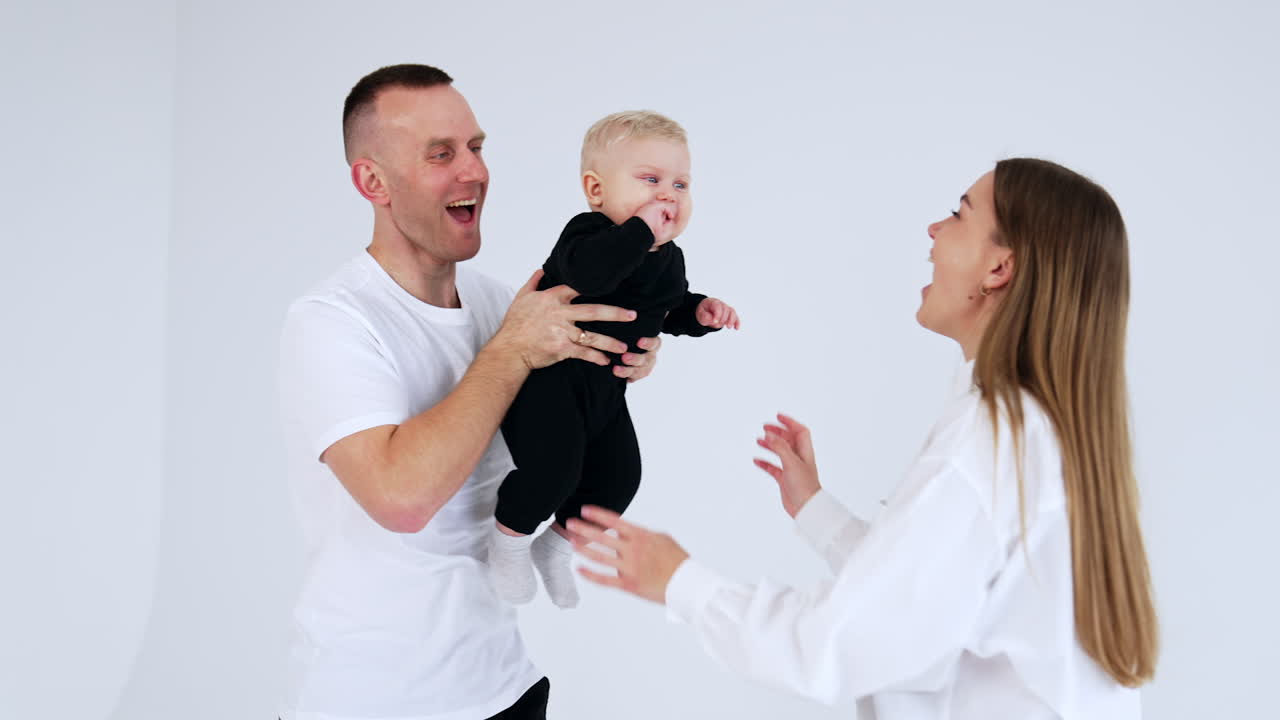 Father holding a cute little baby boy and mother plays with him. Happy parents entertaining their son. White backdrop.