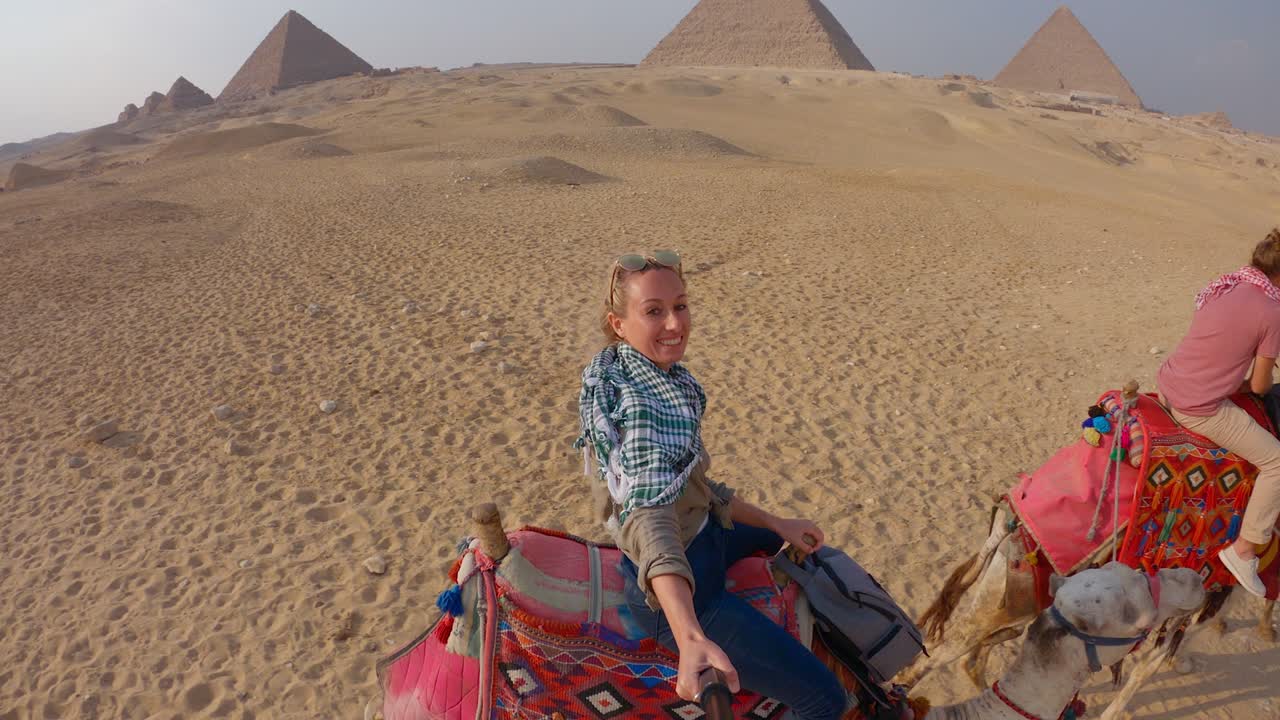 una pareja se toma una selfie montando camellos, las pirámides en el fondo.