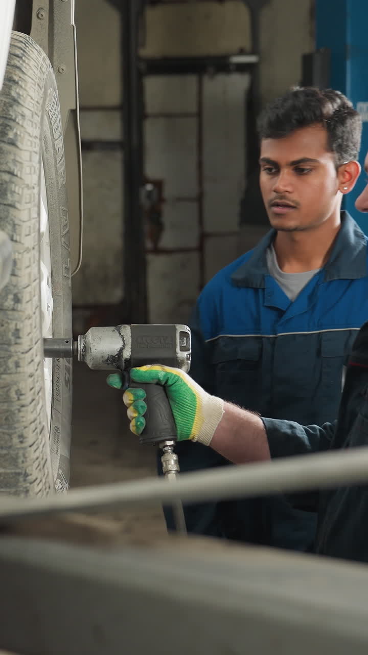 un mecánico en uniforme azul usa una herramienta neumática para apretar los pernos de las ruedas de un coche levantado, mientras que un compañero se acerca, inspecciona el trabajo y asente en aprobación en un taller de automóviles