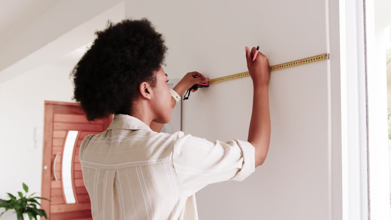 African American woman measuring wall by door for hanging, checking tape and making pencil mark
