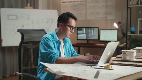 asian man engineer looking at blueprint and typing on a laptop before walking out of the office