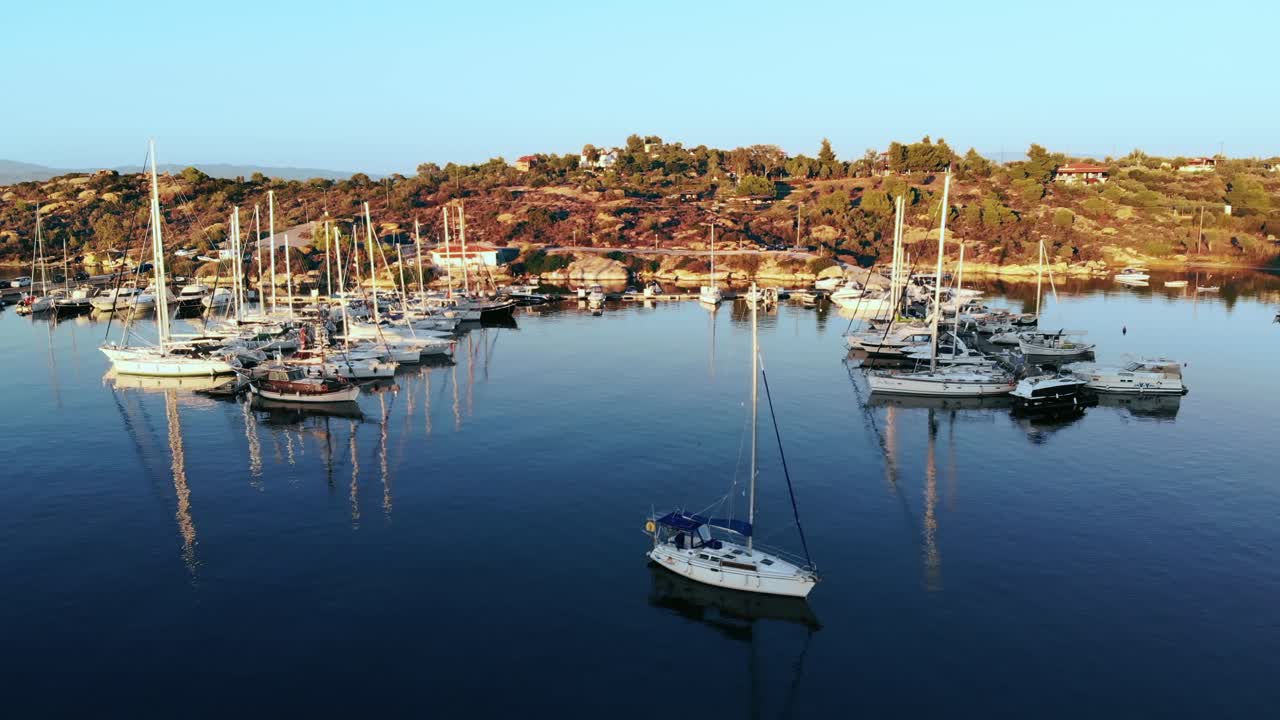 Aerial drone view of Aegean sea port with multiple moored yachts at piers, greenery, blue water, sunset, Greece